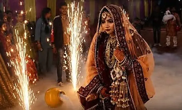 A bride in a red and gold lehenga with a peach dupatta walking between two rows of cold-fire sparklers through low smoke, guests watching from under a marquee.